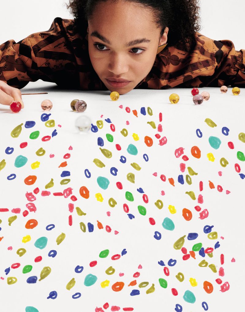 Woman gazes at colorful shapes and marbles scattered across a white surface in a close-up studio shot with shallow depth of field.