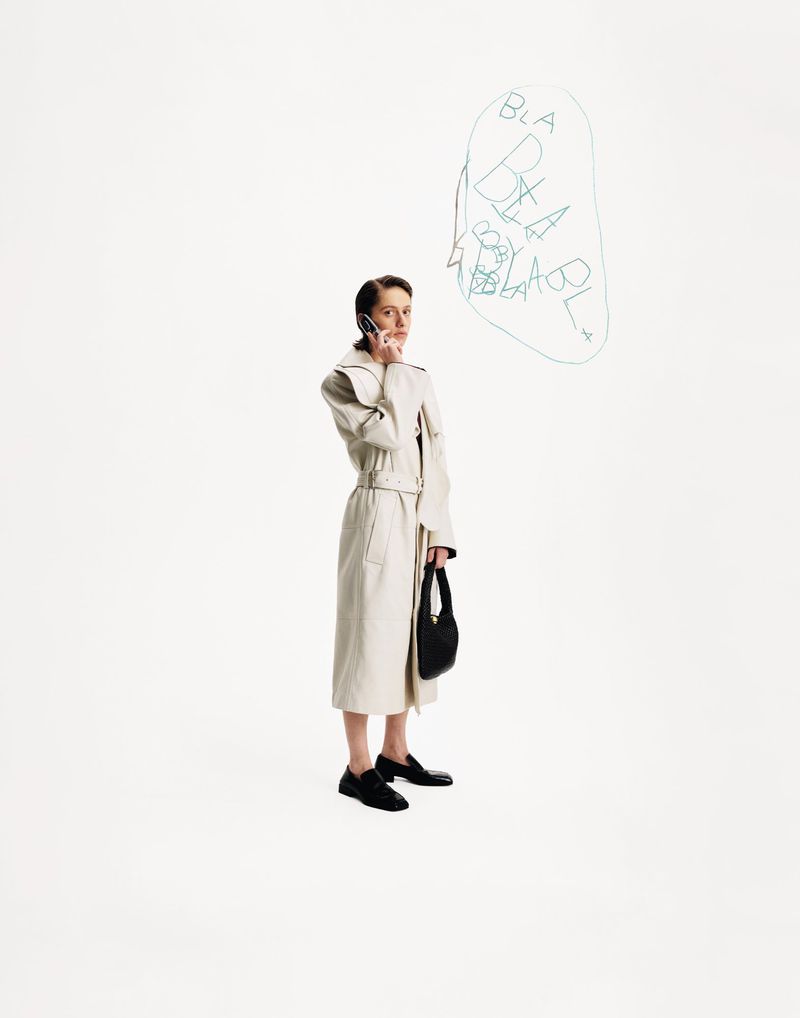 A stylish young woman in a trench coat is holding a phone to her ear while holding a handbag on a clean white backdrop.