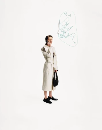 A stylish young woman in a trench coat is holding a phone to her ear while holding a handbag on a clean white backdrop.