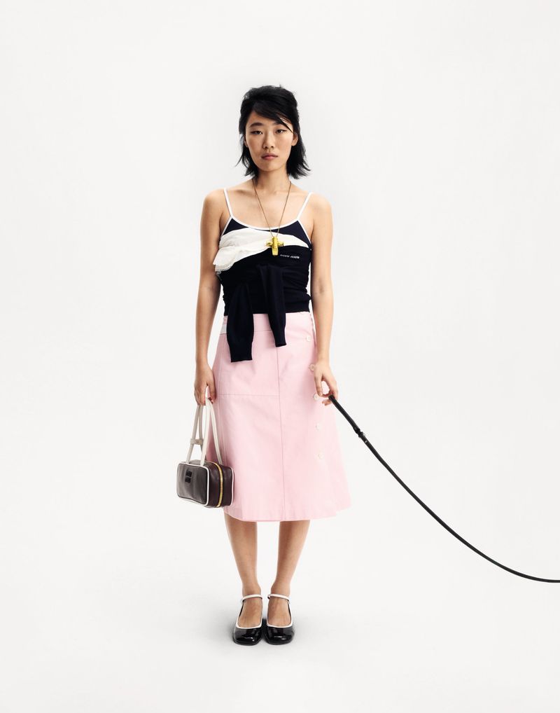 Full shot of an Asian woman in a camisole top, pink skirt and Mary Jane shoes, holding a handbag and a leash, set against a white backdrop.