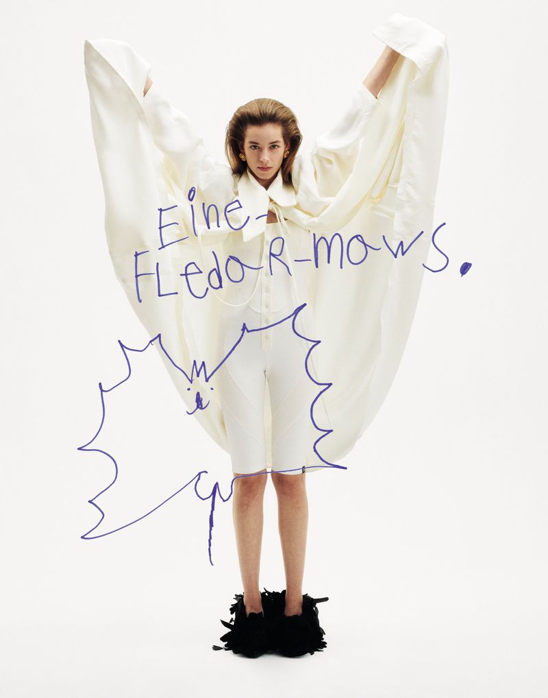 Fashion shot of a woman in white shorts and top with a cape, standing on a black, fringed base, against a stark white background.