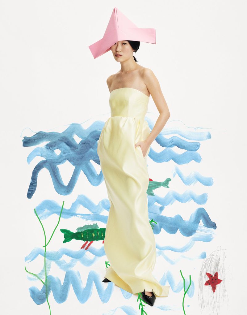 Elegant Asian woman poses in yellow gown and pink hat against a whimsical hand-drawn backdrop of ocean waves and sea creatures.