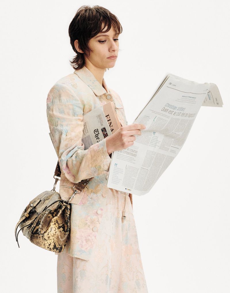 Woman in a pastel floral outfit stands indoors reading a newspaper, with a snakeskin purse, against a bright white neutral background.