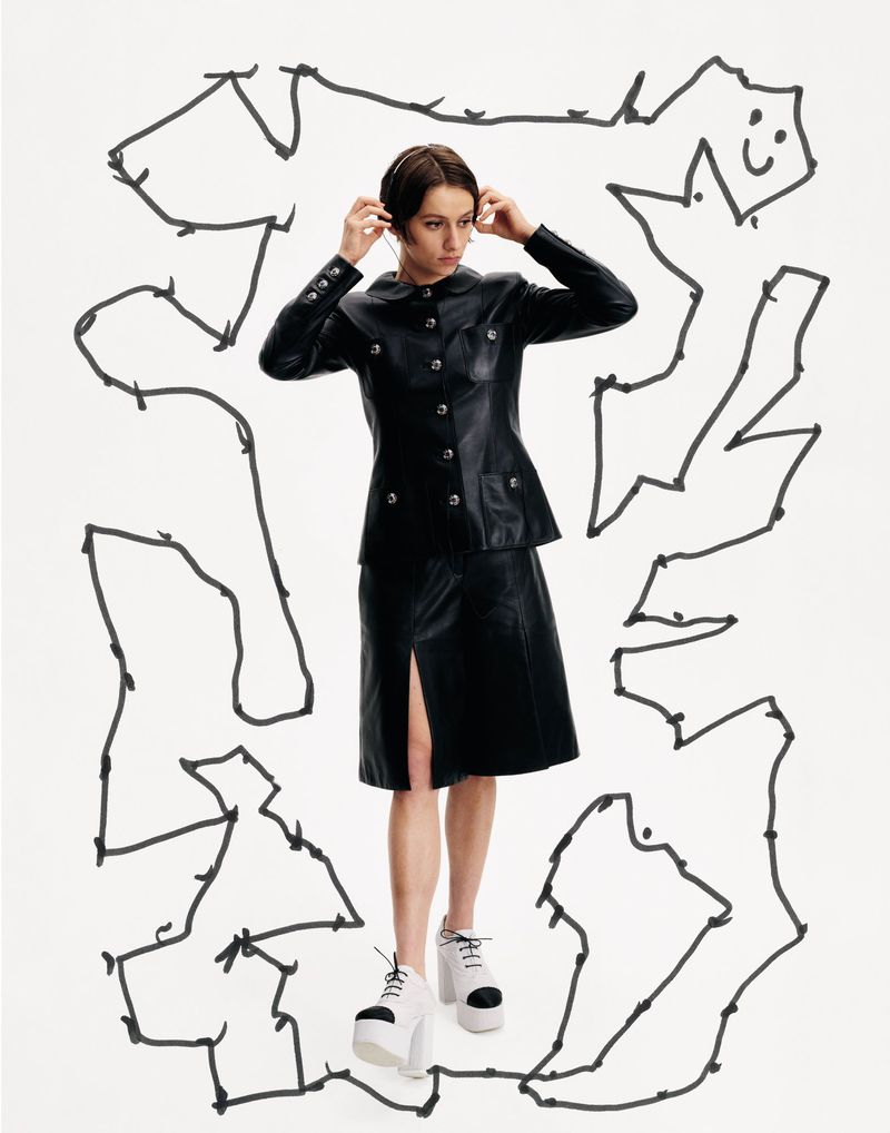 A woman wears a black leather skirt suit with white platform shoes, standing in front of a doodle-covered white background.