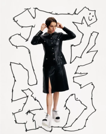 A woman wears a black leather skirt suit with white platform shoes, standing in front of a doodle-covered white background.