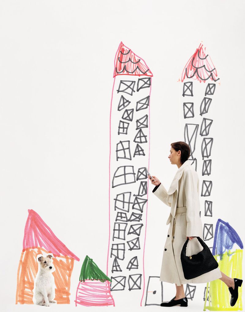 Woman in a trench coat stands near buildings drawn on a white surface with a dog sitting nearby, creating a whimsical urban scene.