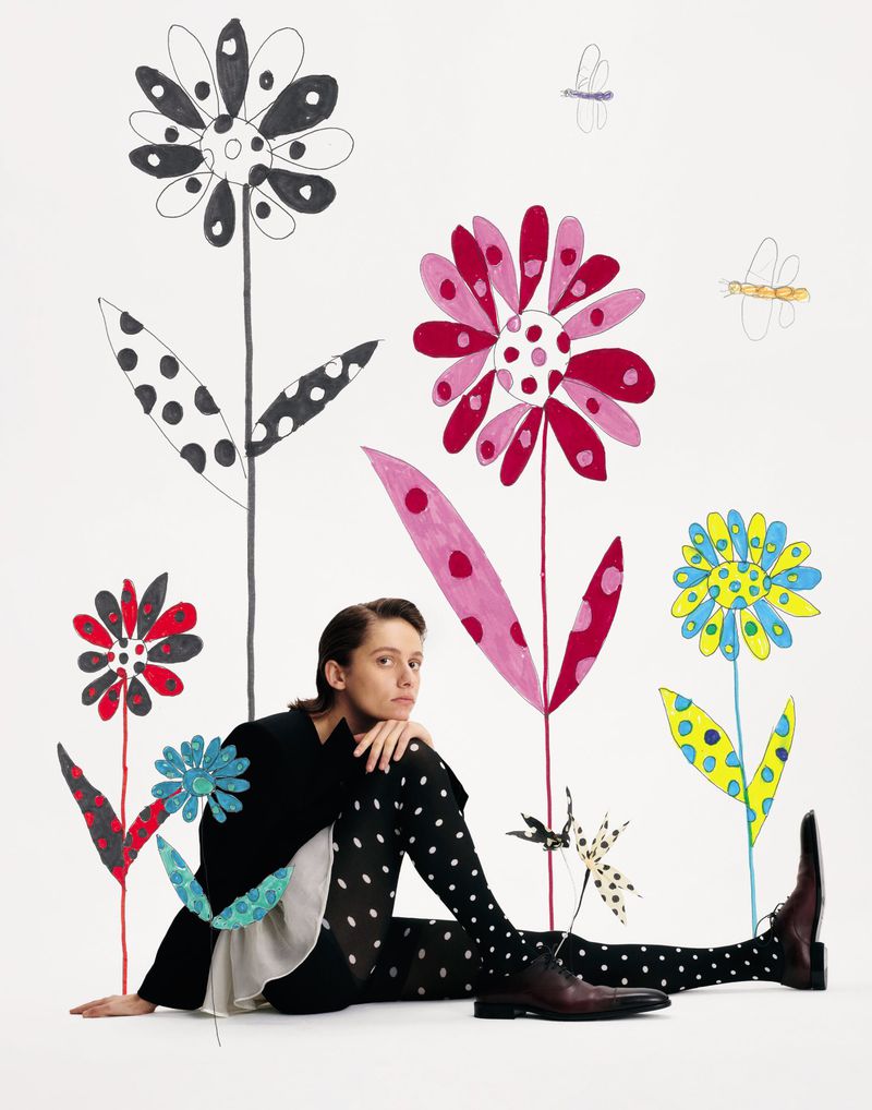 Woman sits in front of a white backdrop with large illustrated polka-dot flowers, wearing polka-dot tights and a black jacket.