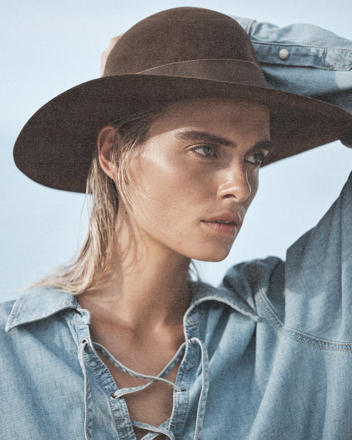 Portrait of a woman wearing a wide-brimmed brown hat and a light blue denim shirt, gazing off to the side with a serene expression.