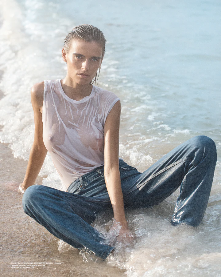 Woman sitting in shallow ocean water wearing a wet white t shirt and blue denim jeans