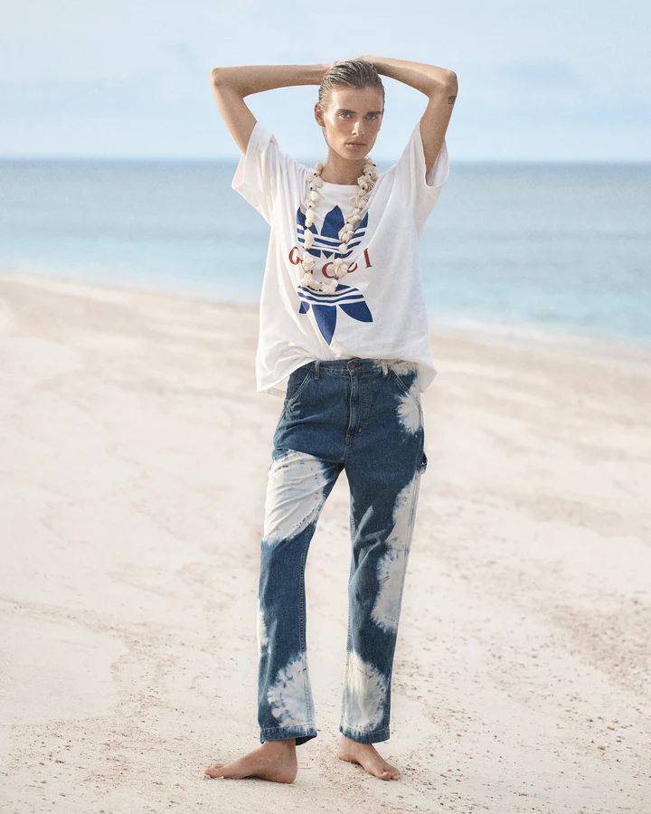 Fashion shot of a young adult woman in a branded t-shirt and tie-dye jeans, posing barefoot on a beach with the ocean in the background.