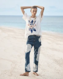 Model standing on a sandy beach wearing a white t-shirt and tie-dye denim jeans with hands behind head.