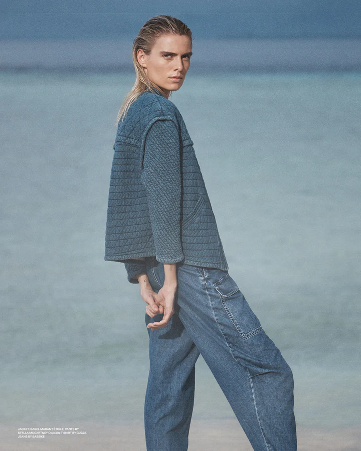 Adult woman poses in a quilted jacket and jeans on a beach with blurred blue ocean and sky as a backdrop, in a fashion editorial shot.