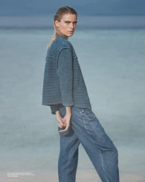 Model with slicked-back blonde hair wearing a textured blue quilted jacket and denim cargo trousers standing by the ocean