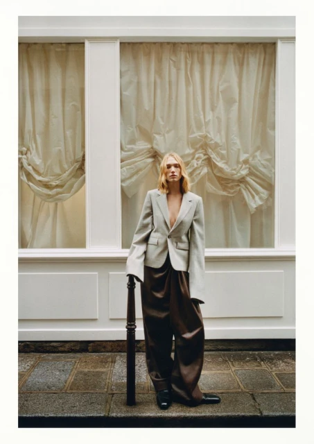 Adult female poses outdoors in an unbuttoned blazer and loose brown pants, beside a white building with decorative curtains and stone sidewalk.
