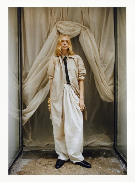 Fashion shot of a fair-skinned adult woman wearing a tan trench coat and white pants, standing indoors against a draped fabric backdrop.