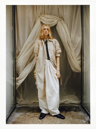 Fashion shot of a fair-skinned adult woman wearing a tan trench coat and white pants, standing indoors against a draped fabric backdrop.