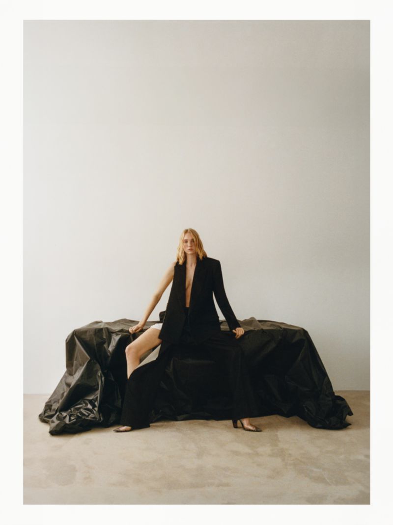A blonde woman sits on a black textile, wearing a black blazer with open front. The setting is a white background, and her legs are crossed.