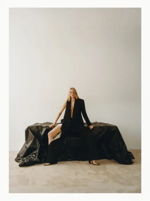A blonde woman sits on a black textile, wearing a black blazer with open front. The setting is a white background, and her legs are crossed.