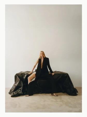 A blonde woman sits on a black textile, wearing a black blazer with open front. The setting is a white background, and her legs are crossed.