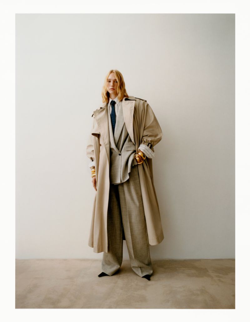 A stylish woman poses indoors wearing a tan trench coat over a gray suit with wide-leg pants and black pointed shoes against a white wall.