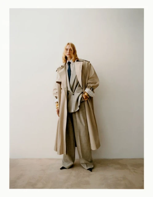 A stylish woman poses indoors wearing a tan trench coat over a gray suit with wide-leg pants and black pointed shoes against a white wall.
