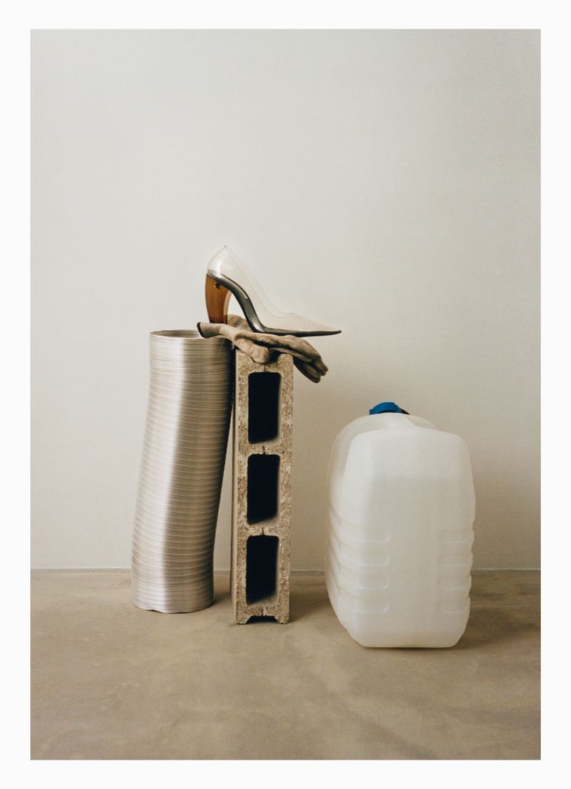 Still life shot in a neutral indoor setting, featuring a high heel shoe atop a cinder block, alongside a ventilation pipe and water container.