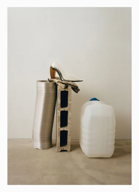 Still life shot in a neutral indoor setting, featuring a high heel shoe atop a cinder block, alongside a ventilation pipe and water container.