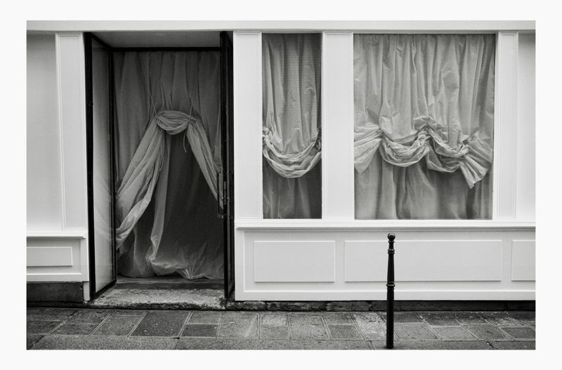 Black and white architectural shot of an open doorway and display windows featuring draped curtains, viewed from a stone sidewalk.
