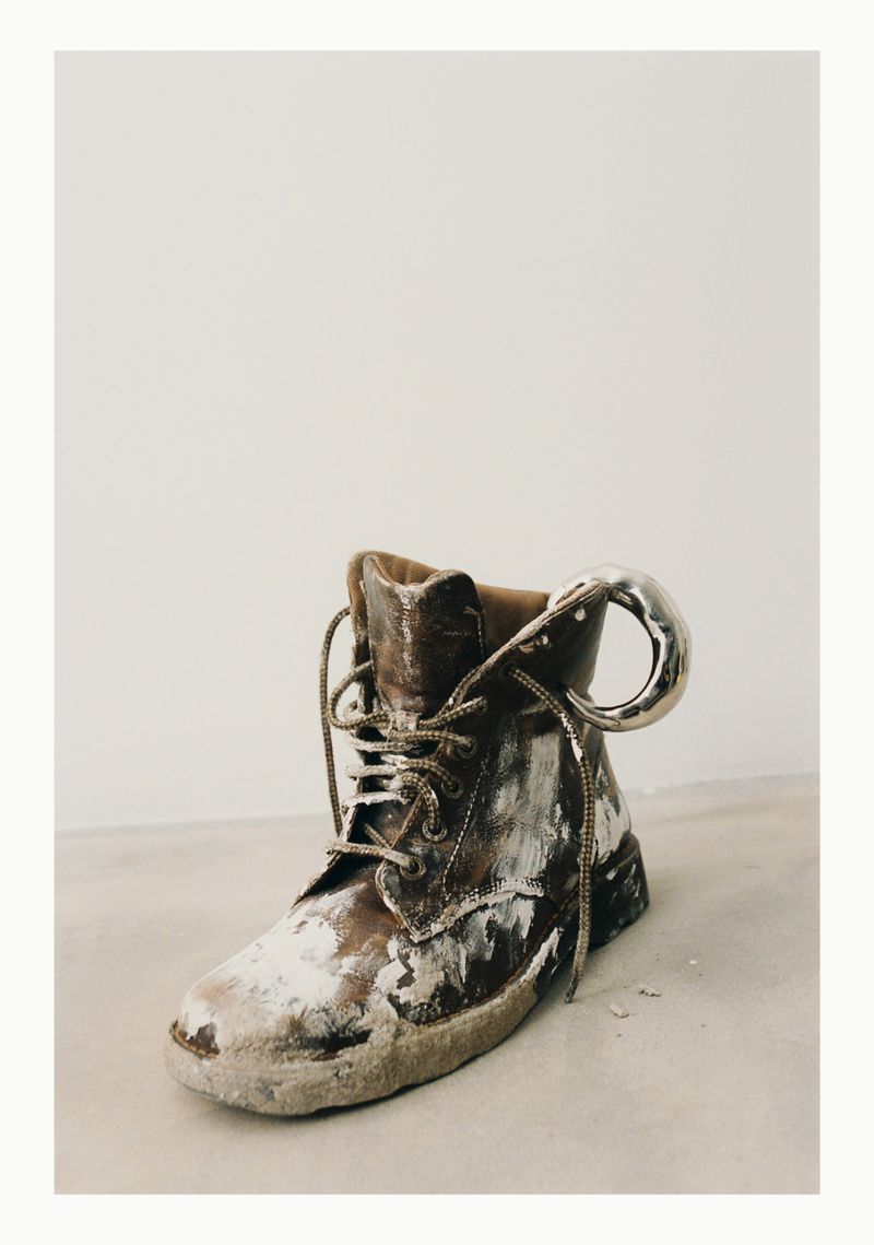Distressed brown leather boot with silver metal pull tab against an off-white background, creating a vintage and artistic composition.