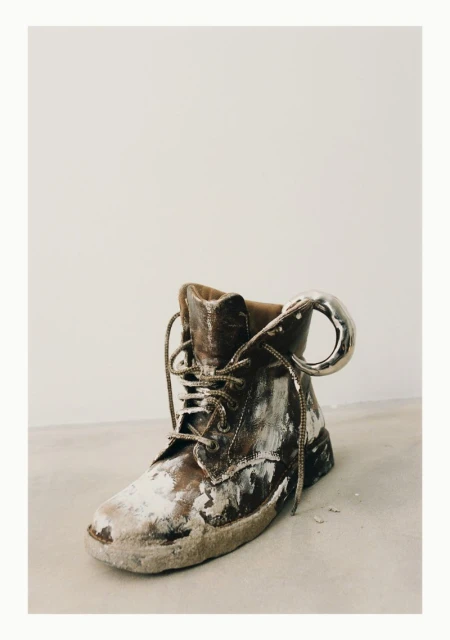 Distressed brown leather boot with silver metal pull tab against an off-white background, creating a vintage and artistic composition.