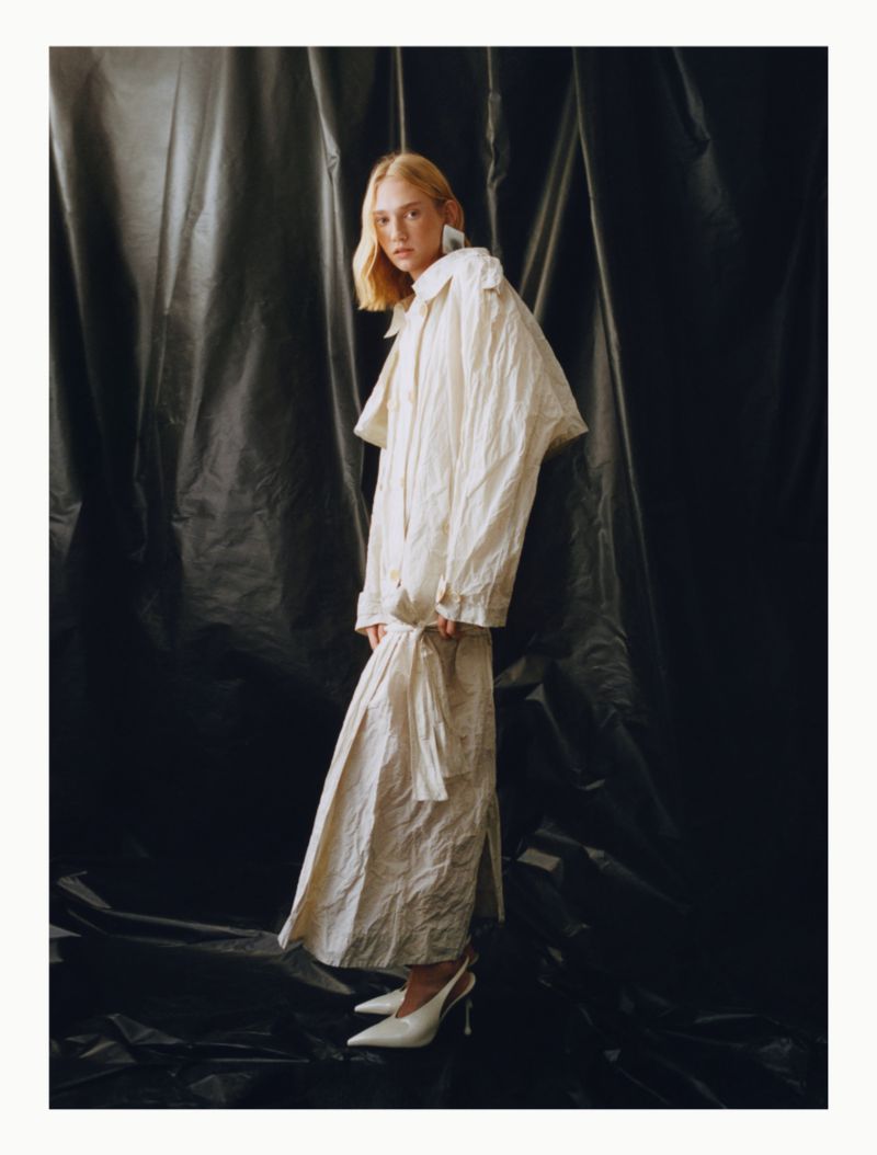 Elegant blonde woman poses in a draped off-white trench coat and matching skirt against a dark, textured backdrop in a fashion-forward shot.