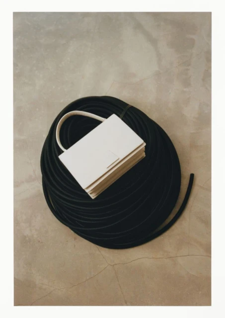 Elevated shot of a light-colored rectangular handbag with a handle resting on a coil of black tubing against a neutral background.