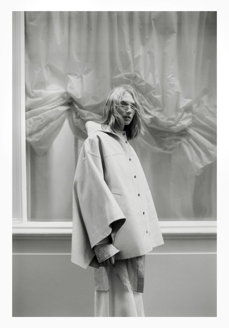Monochrome fashion shot of a woman in an oversized jacket in front of a window with draped curtains in an indoor setting.