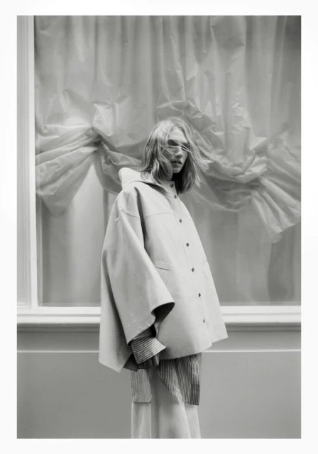 Monochrome fashion shot of a woman in an oversized jacket in front of a window with draped curtains in an indoor setting.