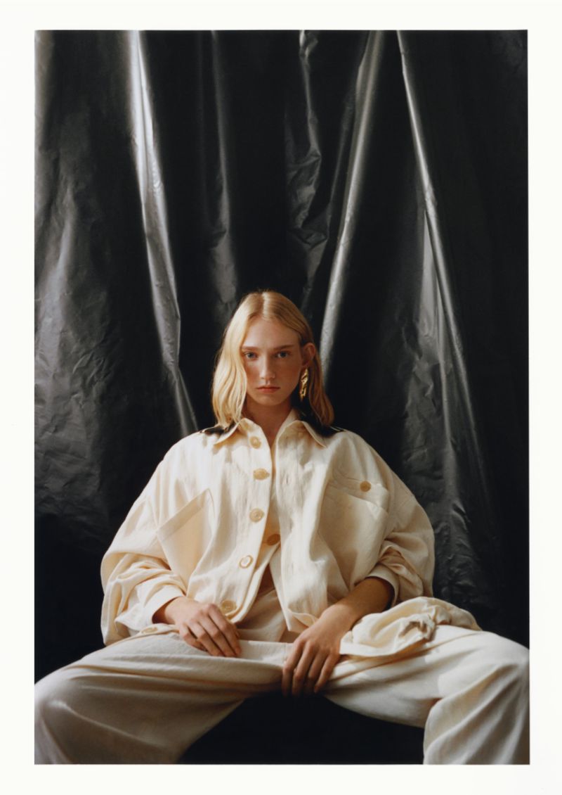 Fashion shot of a blonde adult woman wearing a cream-colored jacket and pants, sitting against a dark, draped fabric backdrop.
