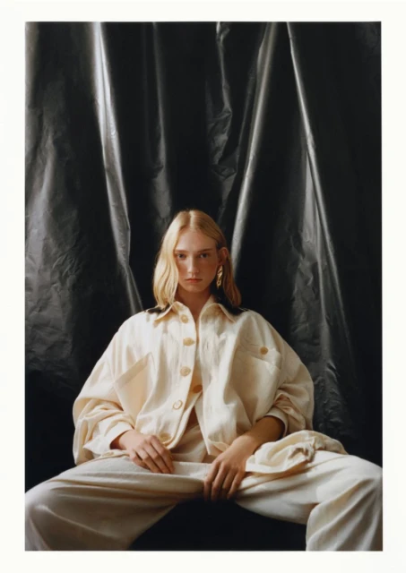 Fashion shot of a blonde adult woman wearing a cream-colored jacket and pants, sitting against a dark, draped fabric backdrop.