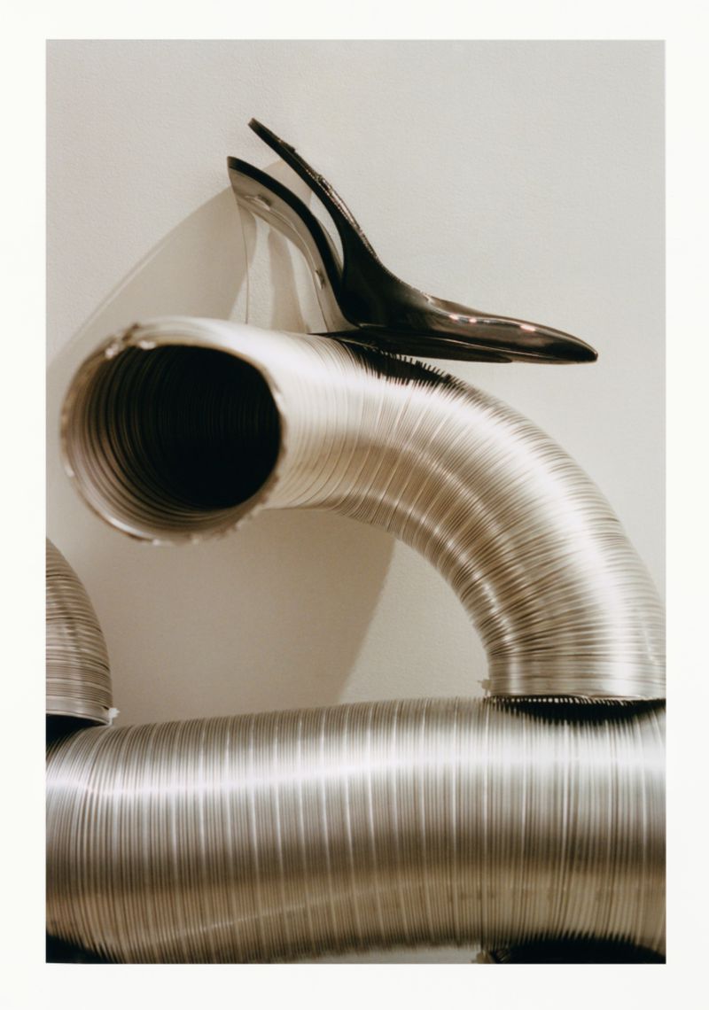 Still-life photograph features industrial aluminum ventilation tubes and a black high heel shoe, creating a stark, high-contrast composition.