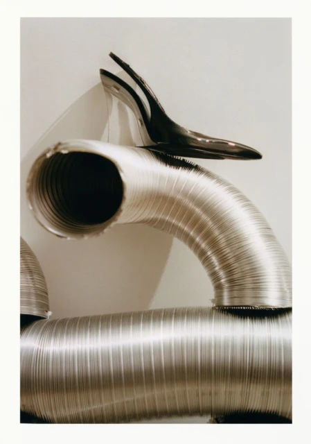 Still-life photograph features industrial aluminum ventilation tubes and a black high heel shoe, creating a stark, high-contrast composition.