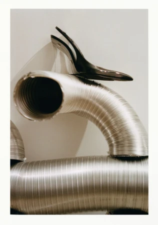 Still-life photograph features industrial aluminum ventilation tubes and a black high heel shoe, creating a stark, high-contrast composition.