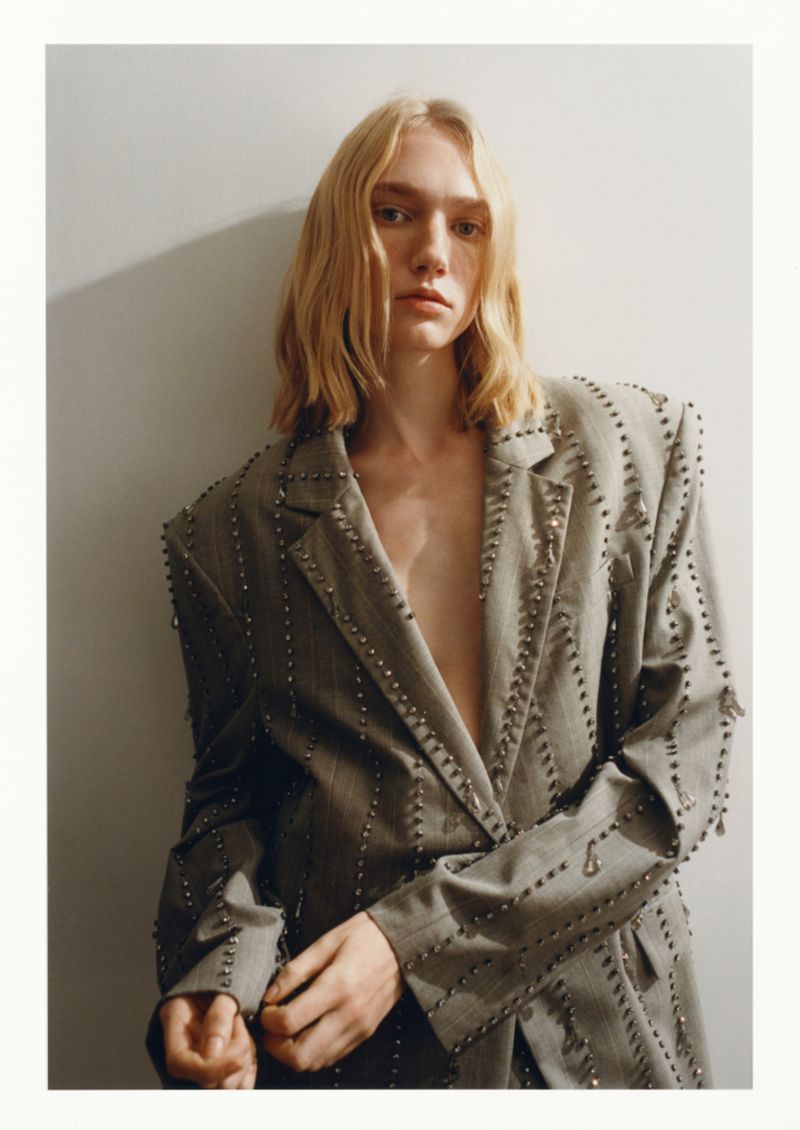 Woman with blonde hair poses in an oversized grey blazer with silver embellishments against a plain grey wall in a studio setting.
