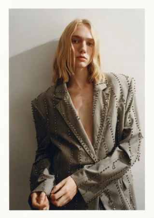 Woman with blonde hair poses in an oversized grey blazer with silver embellishments against a plain grey wall in a studio setting.