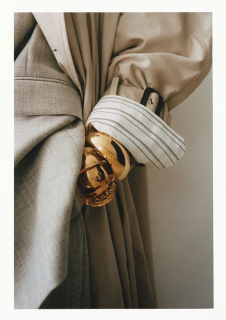 Close-up studio shot of two coats, one light tan and one light grey, draped together with a gold bracelet.