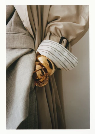 Close-up studio shot of two coats, one light tan and one light grey, draped together with a gold bracelet.