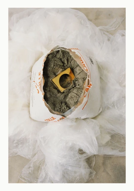 White plastic bag filled with grey rubble and square gold ring in the middle, surrounded by a translucent white plastic sheet.