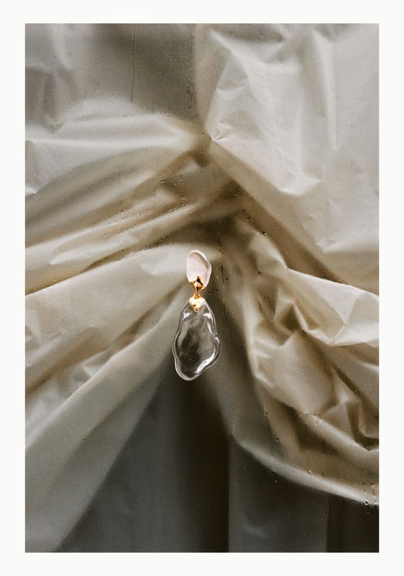 Close-up studio shot of a translucent sculptural earring with a gold connector, set against a light beige, wrinkled fabric background.