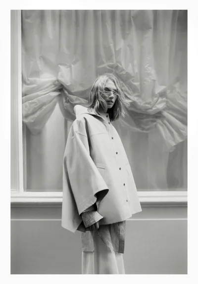 Model wearing a buttoned cape and striped shirt standing in front of crinkled draped fabric for Harper's Bazaar Italia