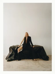 Model seated on a black draped structure against a plain white studio wall wearing a black blazer and trousers
