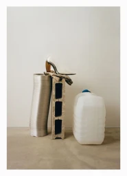 High-heeled shoe resting on a concrete cinder block beside a metal duct and plastic jug on a concrete floor