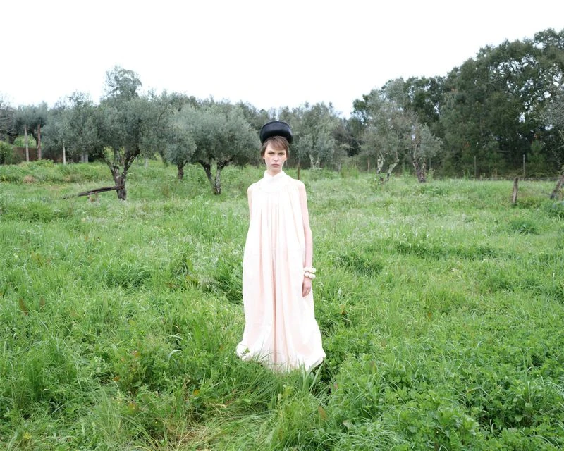 Woman in long pink dress stands in grassy field with trees under an overcast sky, accessorized with a black hat and white bracelets.