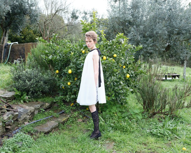 A fair-skinned young adult female in a white dress with black accents stands in a grassy field near a lemon tree on a sunny day.